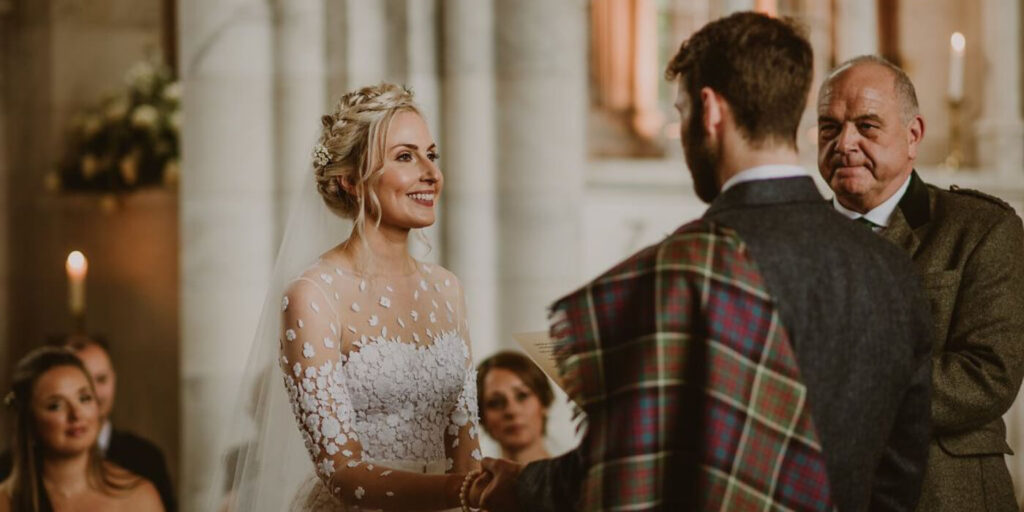 Bride wearing wedding dress holding hands of groom who is wearing Scottish tartan at their wedding ceremony