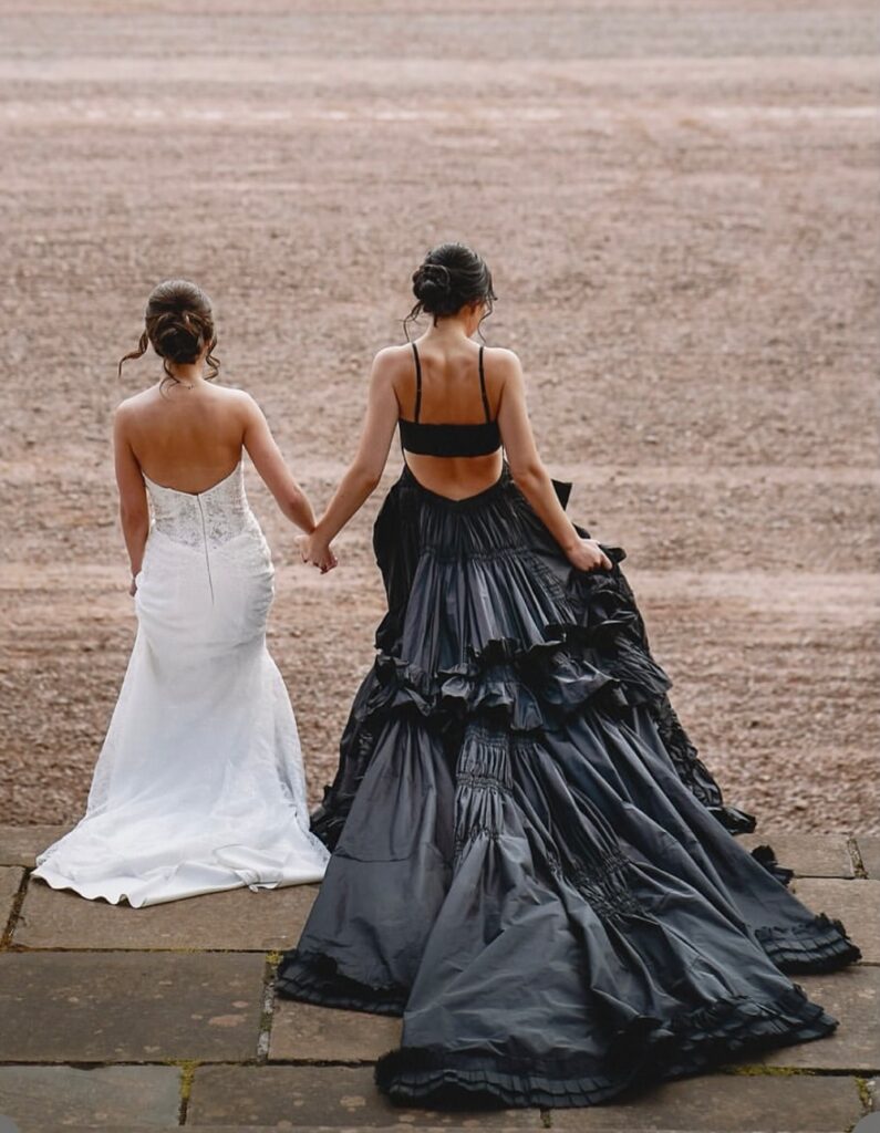 Two brides walking away holding hands on a beach. One bride wears a white dress and one wears a black dress. 