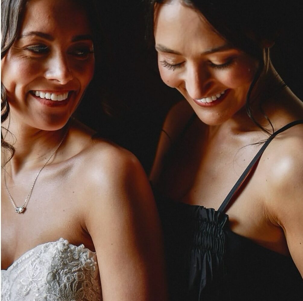 Same sex couple Fiona and Sasha, one bride in a white dress and another in a black dress, smiling at each other.
